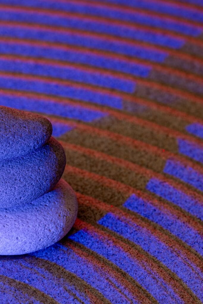 Stacked zen stones on grooved sand illuminated by blue and red light, creating a serene ambiance.